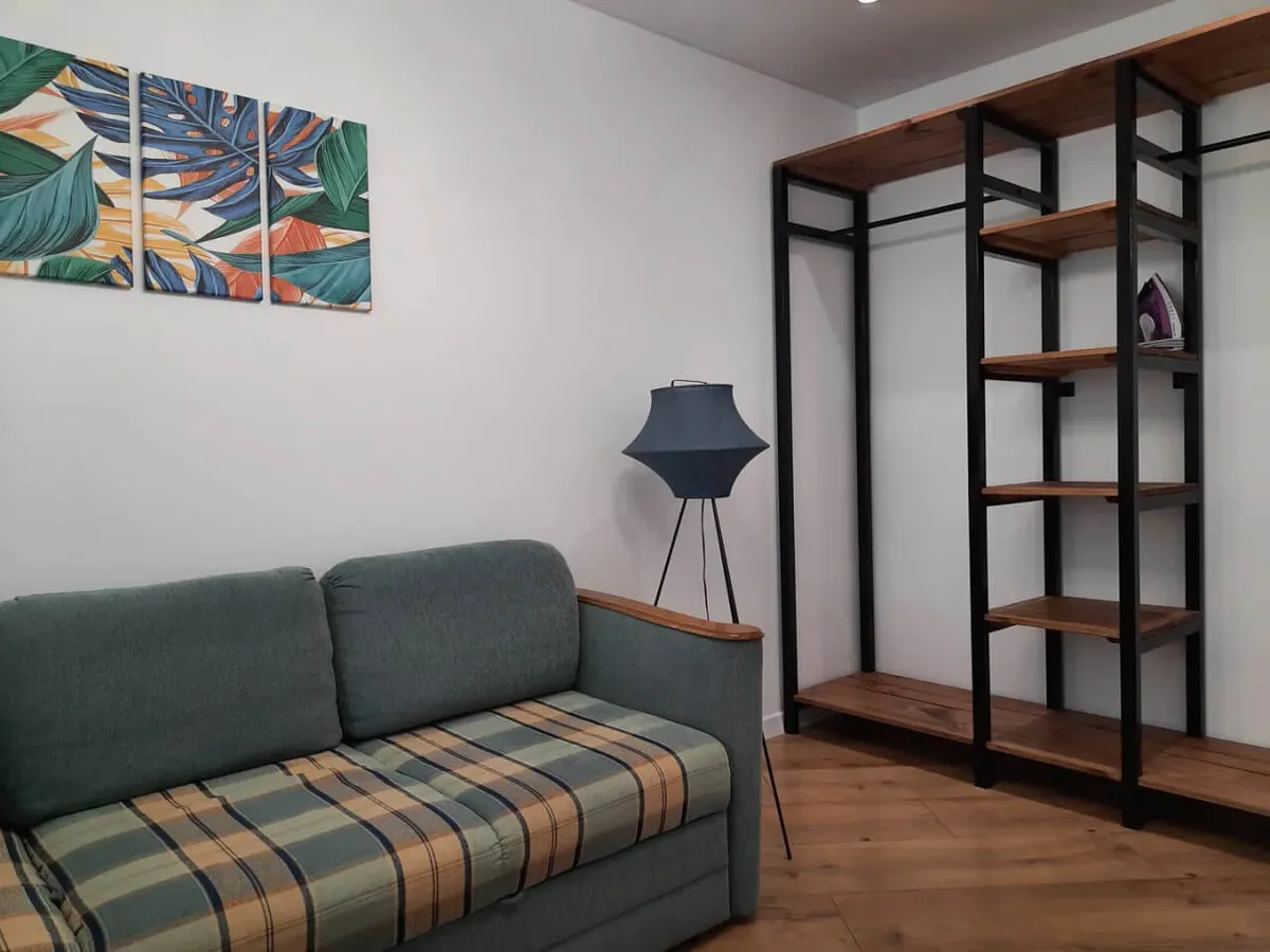 The image showcases a bright, stylish living area with a greenish-gray sofa adorned with a plaid cushion, set against a backdrop of vibrant tropical leaf triptych wall art. A modern tripod floor lamp with a blue shade adds a touch of elegance, while a wooden and black metal shelving unit provides both storage and homey organization. The overall feel is inviting, practical, and comfortable—ideal for relaxing or working during your stay.