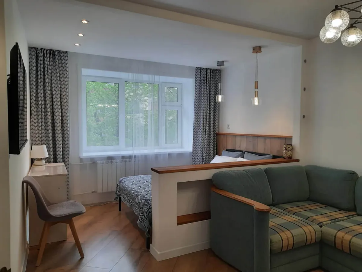This image displays the main living area, featuring a green cushioned sofa with plaid pattern, a bed area partially separated by a half wall with a wooden headboard, a desk and chair near the window, and large windows with patterned curtains and sheer drapes. The space is filled with natural light, and the layout encourages both relaxation and productivity.