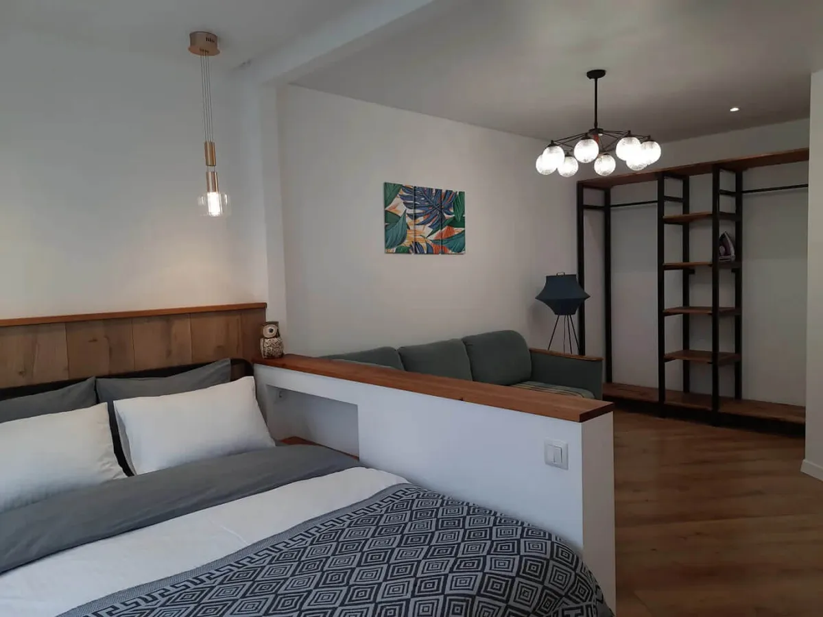 A view of the living area with a bed dressed in patterned blankets and pillows, a green sofa behind a partial wall, open wardrobe with wooden shelves, and modern ceiling lights. The space is inviting and functional, with clear separation of lounge and rest areas, and is designed for both relaxation and light productivity.