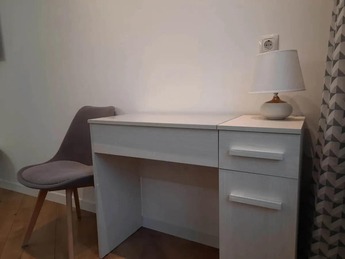 A quiet corner with a small white desk, gray cushioned chair, table lamp, and geometric patterned curtain. The setup is tailored for focused activities such as working on a laptop, reading, or organizing small tasks, and is softly lit for comfort.