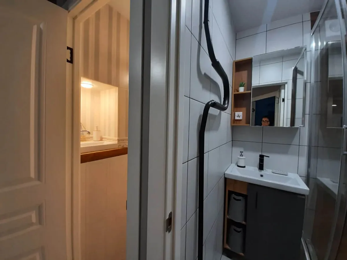 A compact yet well-equipped bathroom with a modern sink and mirror, a towel warmer on the wall, a glass-enclosed shower, and soft lighting from the adjacent kitchen area. The space is clean, practical, and designed for comfort and efficiency.