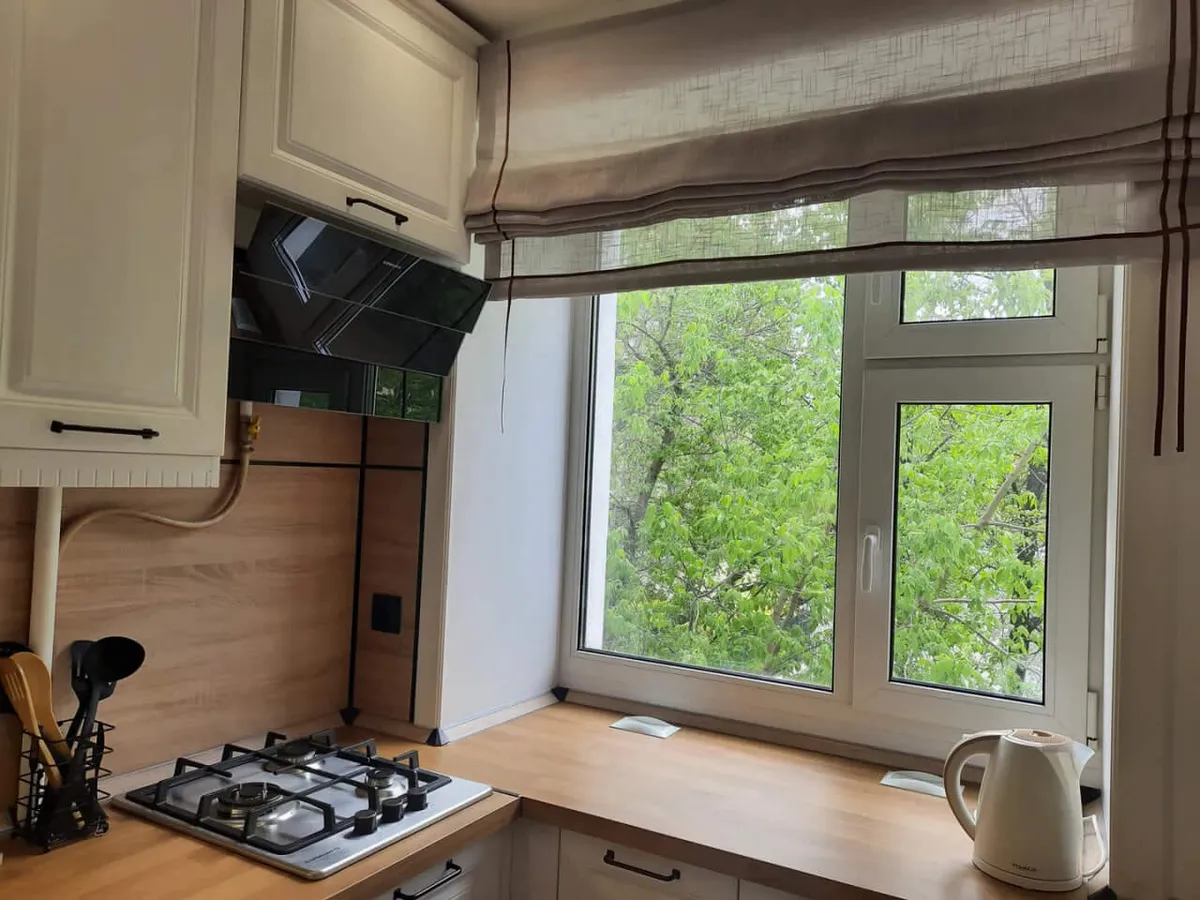 A peaceful kitchen scene featuring a gas stove with cooking utensils, a large window overlooking green trees, a wooden countertop, and an electric kettle. Natural light floods the space, highlighting the kitchen's functionality and inviting atmosphere.
