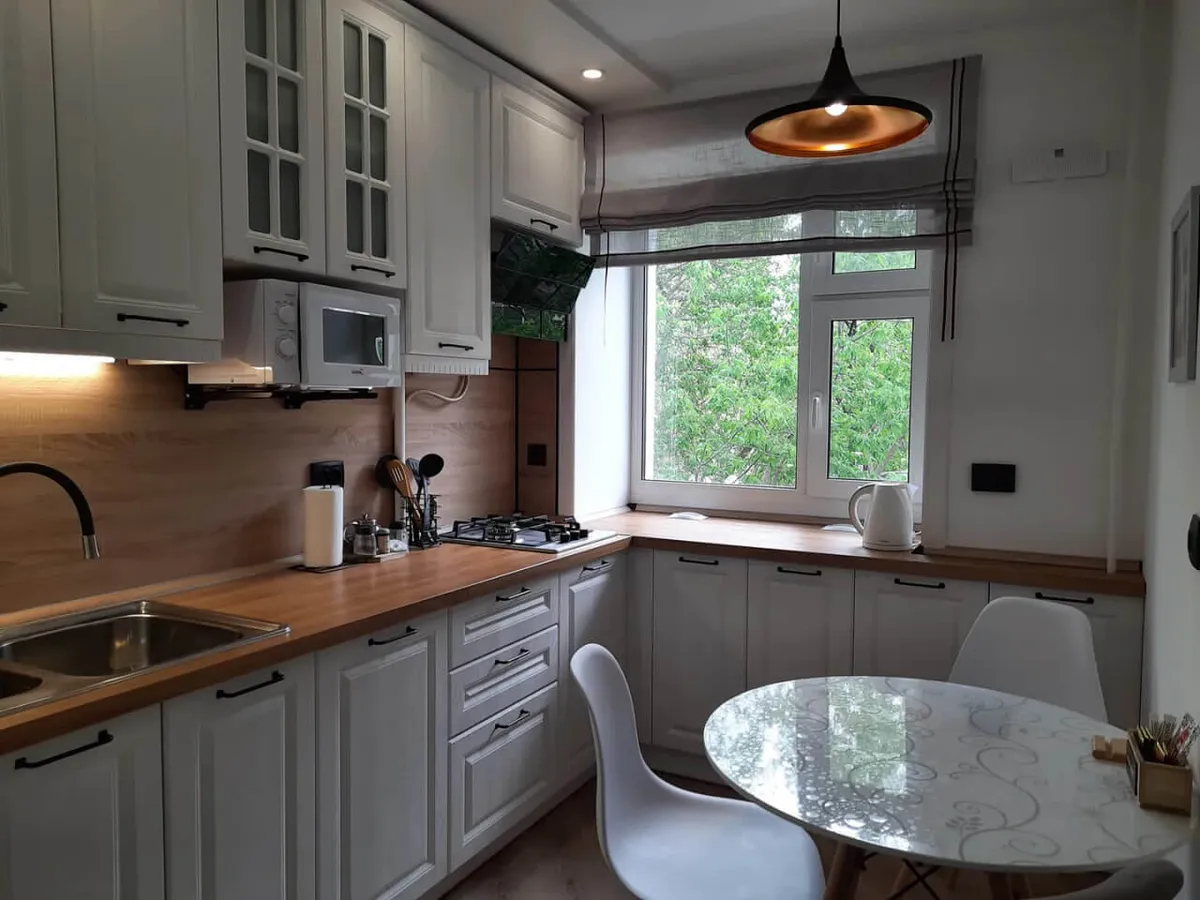 A bright, inviting kitchen with a wood-toned countertop featuring a built-in stove, a large window offering views of greenery, a round glass-topped dining table, and white cabinetry with black handles and a built-in microwave. The scene is tidy and highlights both the kitchen and dining area, suggesting plenty of space for cooking and enjoying meals.