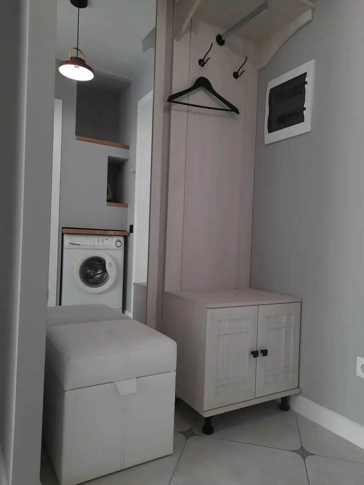 This image showcases a compact, well-organized utility nook featuring a white washing machine, a small white storage cabinet for supplies, black wall hooks with a hanger, and a white cushioned ottoman. The setting is orderly, practical, and clearly designed for everyday laundry tasks and efficient storage.