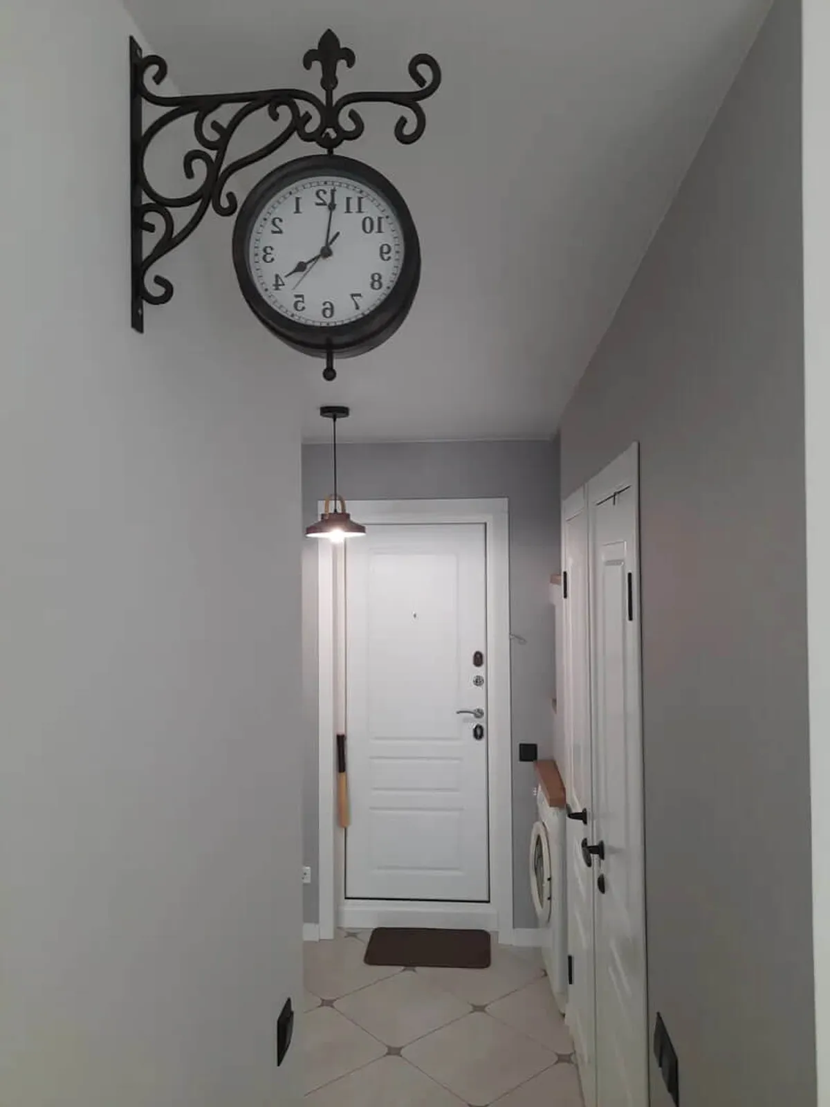 This image shows the apartment entryway with a decorative vintage-style wall clock, a modern white front door, and the washing machine partially visible behind the door. A pendant light brightens the space, and the scene suggests ease of arrival and access to laundry facilities.