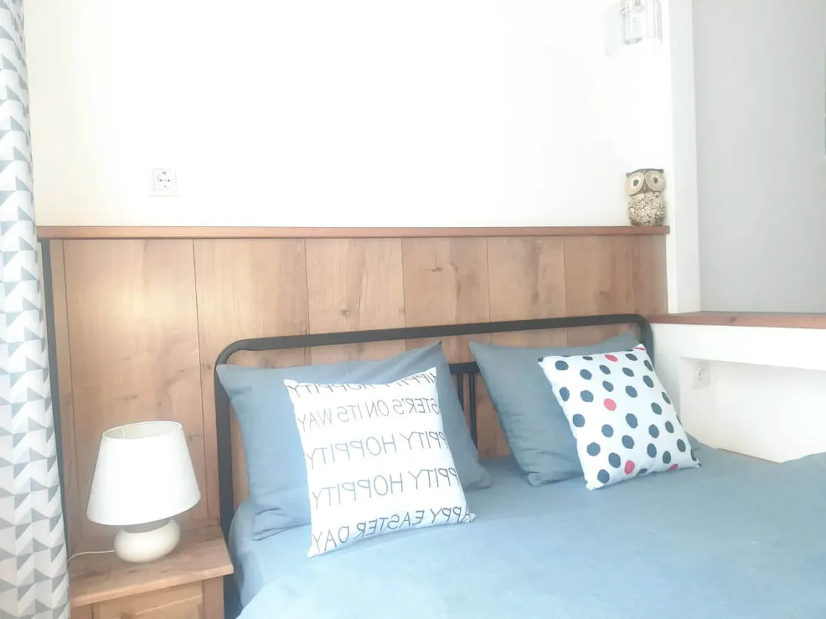 A cozy bedroom featuring a neatly made bed adorned with decorative pillows, a wooden headboard, a white bedside lamp, a small side table, and a charming owl figurine on a shelf. The space exudes warmth and comfort, inviting relaxation and restful sleep.